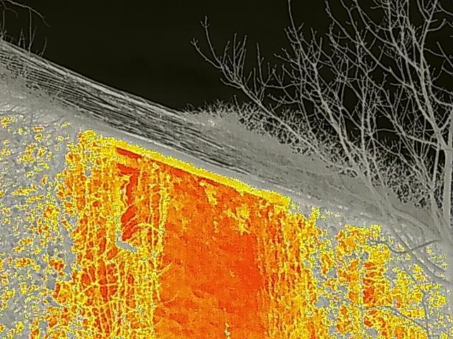 A thermal imaging photograph showing the roof and wall section of a building with heat signatures in orange and yellow.