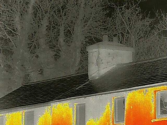 Thermal imaging photograph showing the roofline, chimney, and upper walls of a stone building with trees in the background.