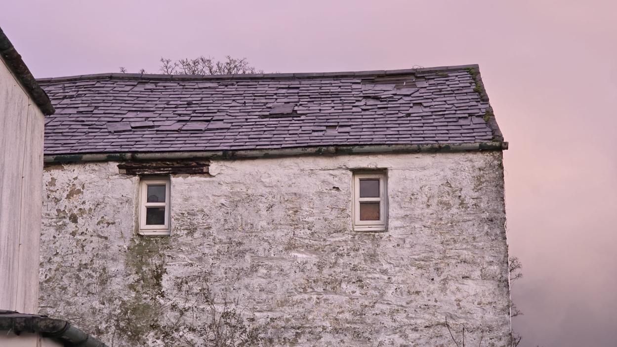 A photograph showing the upper gable end of an old white-washed stone building with a slate roof and two small windows.