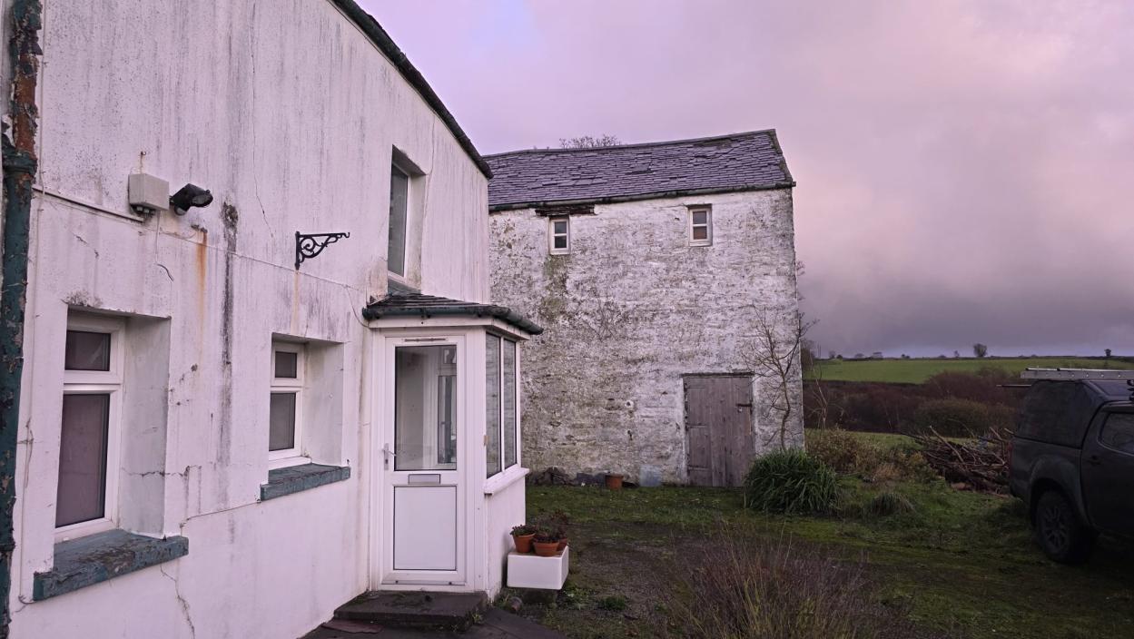 Exterior photograph showing a white-rendered house and an adjacent stone barn or outbuilding in a rural setting.