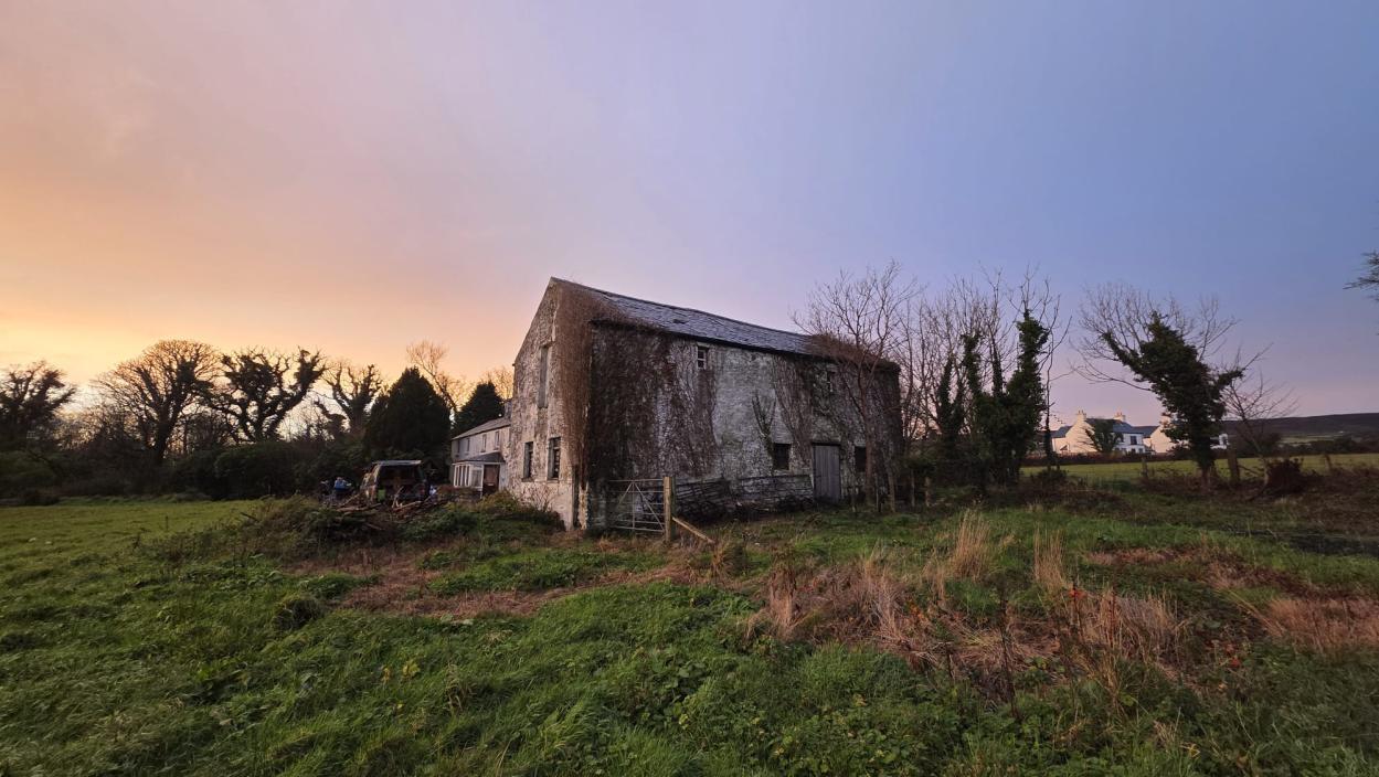 A photograph showing a large, weathered stone barn situated in a rural field at dusk.