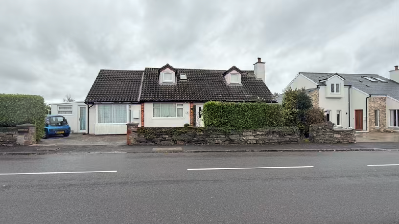 Street-level photograph of a detached bungalow with a dark tiled roof and dormer windows. A white single-story lean-to extension is visible on the left side, fronted by a stone wall and driveway.