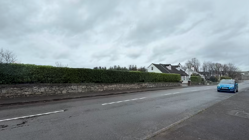 A street-level photograph showing a road with a stone wall and hedge on the left, and white houses in the background under an overcast sky.