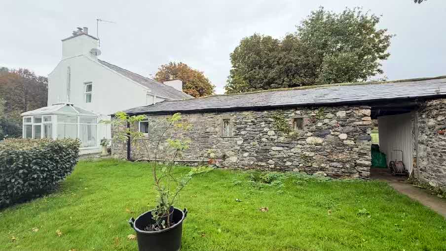 A photograph showing a white house with a glass sunroom attached to the side, adjacent to a long stone outbuilding with a slate roof and a grassy yard.