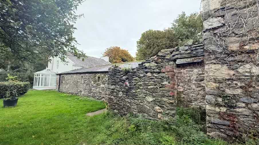 A photograph showing a grassy garden area with a white house featuring a conservatory/sunroom in the background and a crumbling stone wall in the foreground.