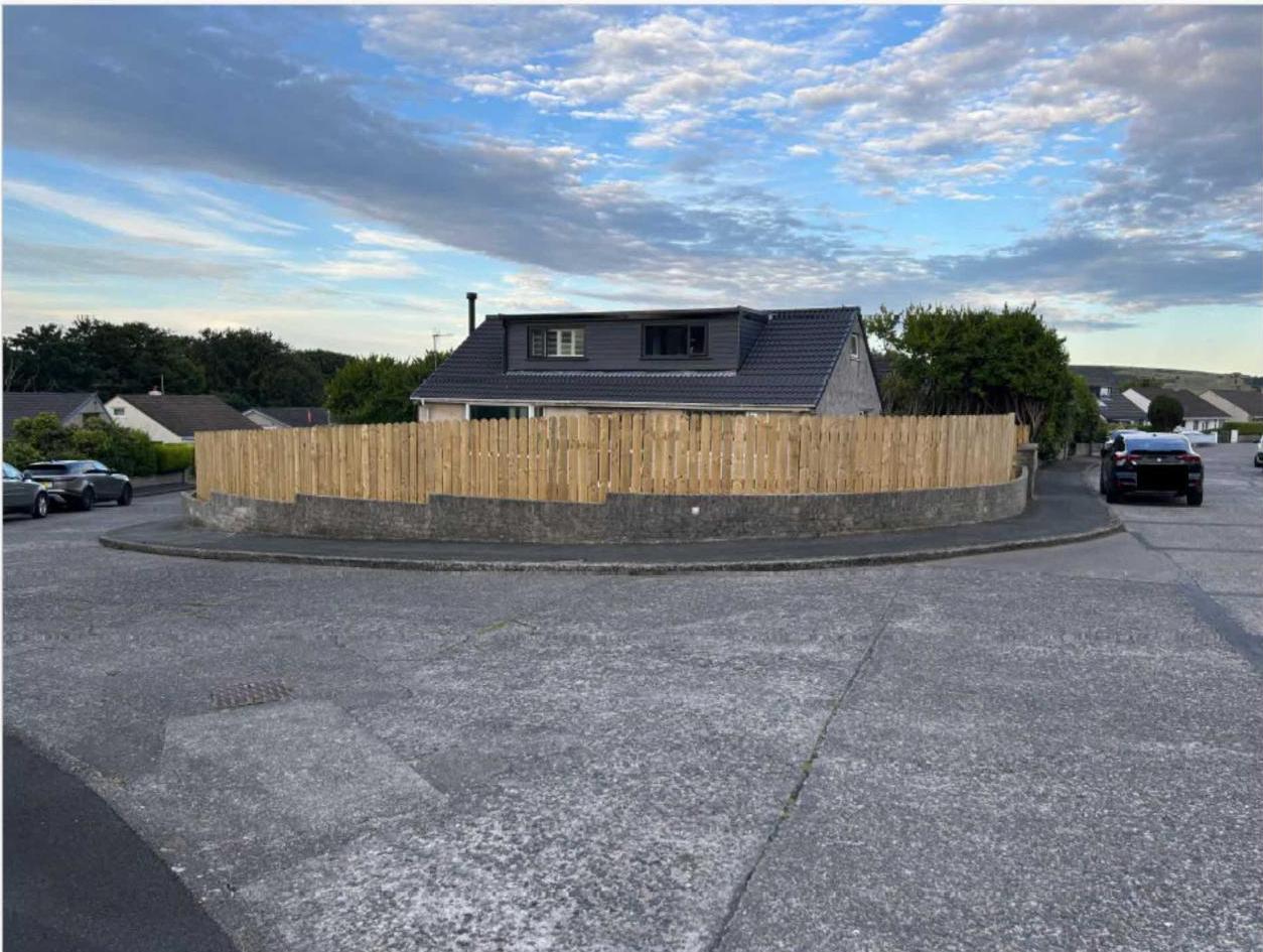 A photograph showing a residential property with a newly installed wooden fence on a stone retaining wall at a street corner. The foreground features a paved hardstanding area with a house visible behind the boundary.