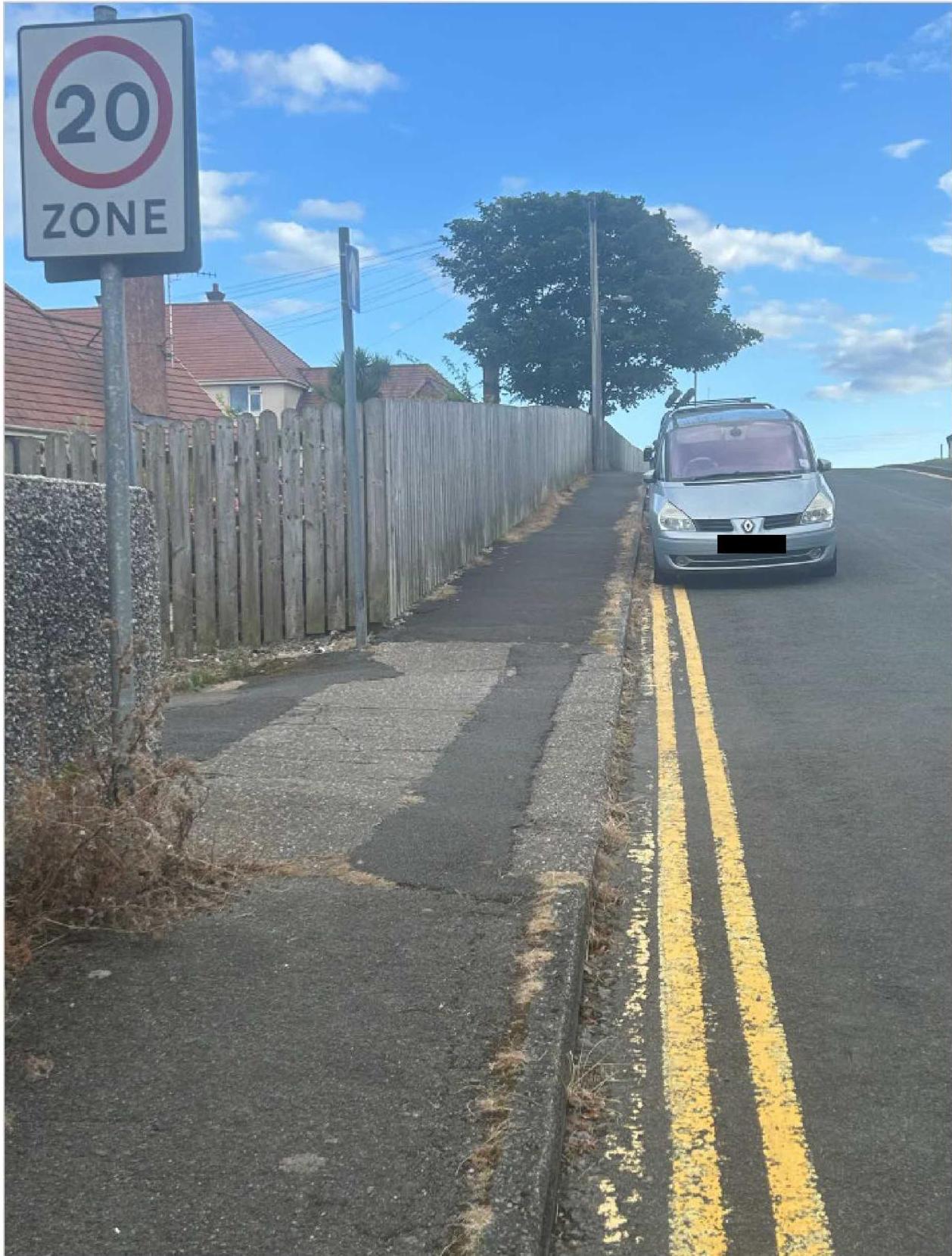 A street-level photograph showing a wooden fence running along a property boundary next to a paved hardstanding area. A silver van is parked on the adjacent road with double yellow lines, and a '20 ZONE' sign is visib...