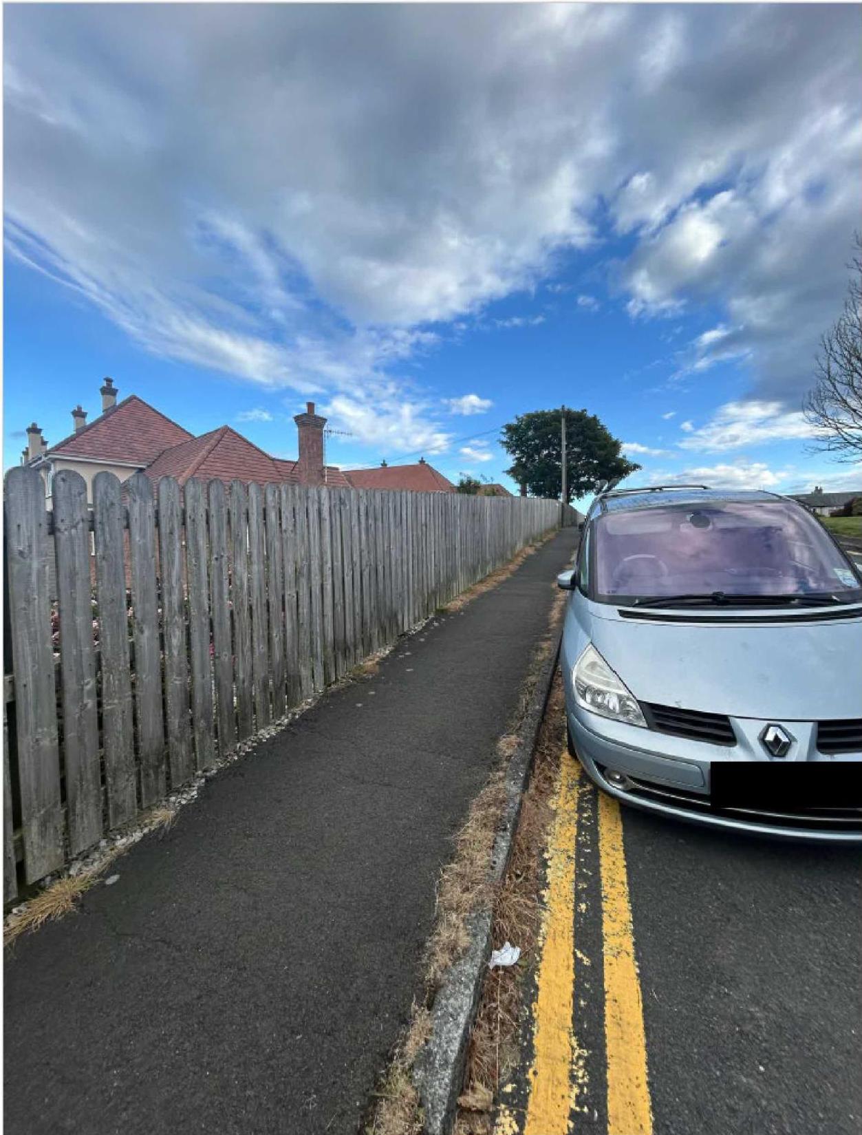 A photograph showing a weathered wooden fence running alongside a paved path and road, with a silver car parked nearby and residential houses visible in the background.