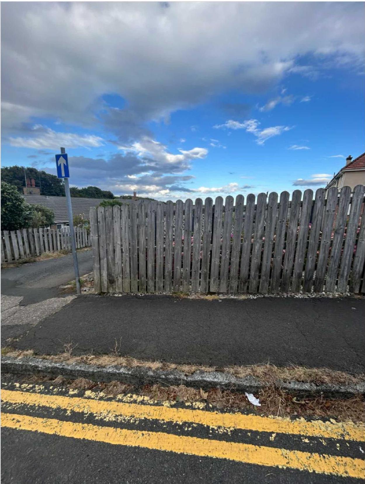 A photograph showing a wooden picket fence and a section of asphalt hardstanding adjacent to a road with double yellow lines.