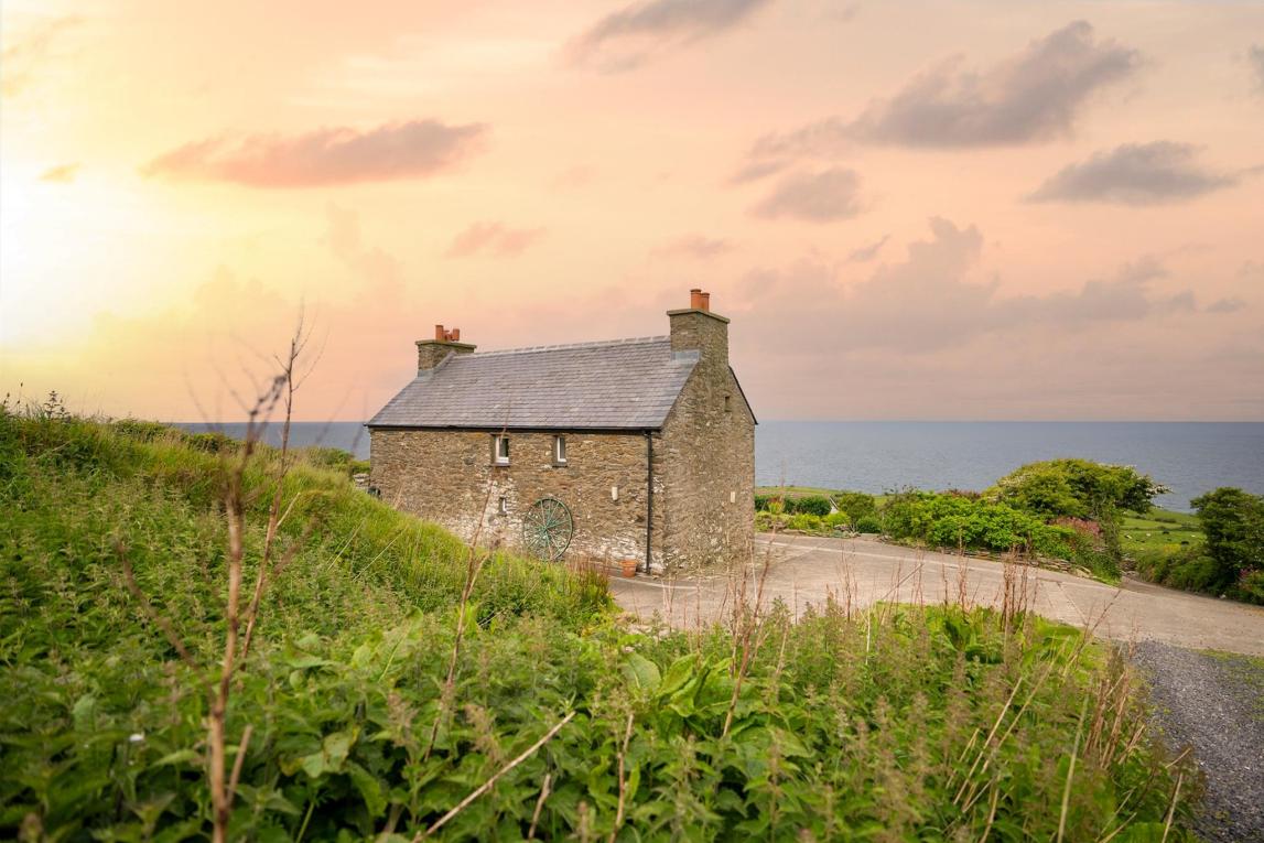 A scenic photograph of a stone building situated on a grassy slope overlooking the sea at sunset.