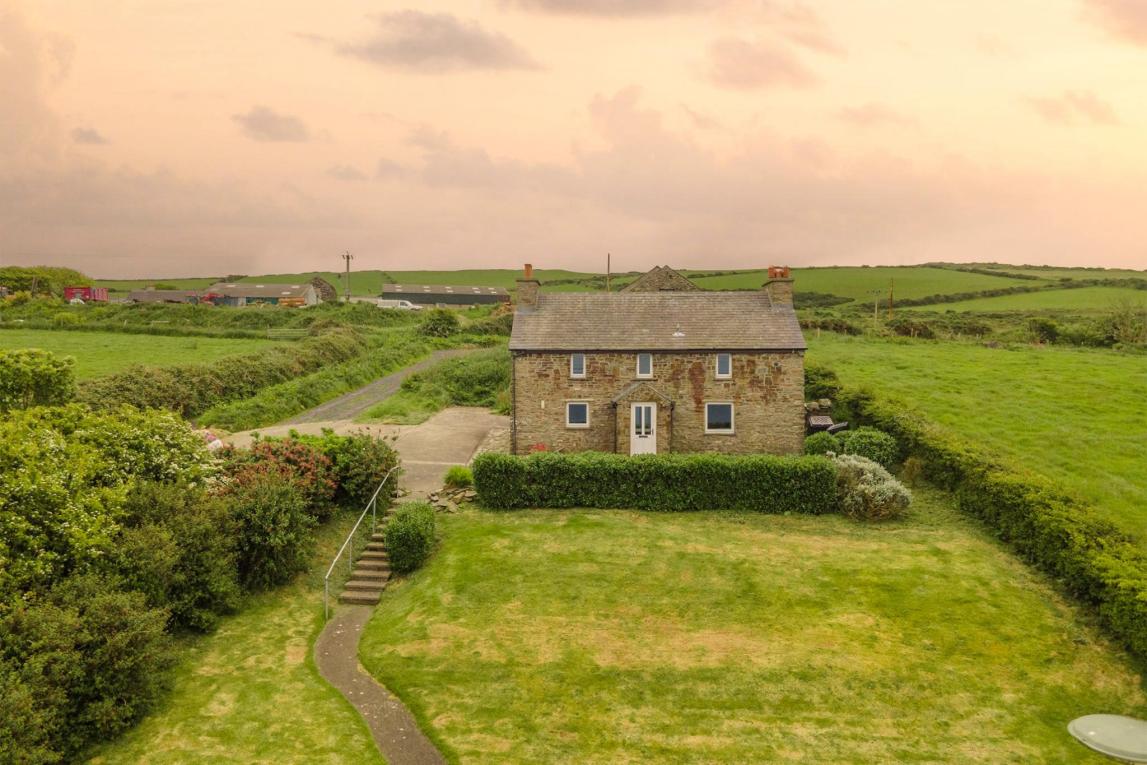 A photograph showing a detached stone house situated in a rural landscape with green fields and a driveway.