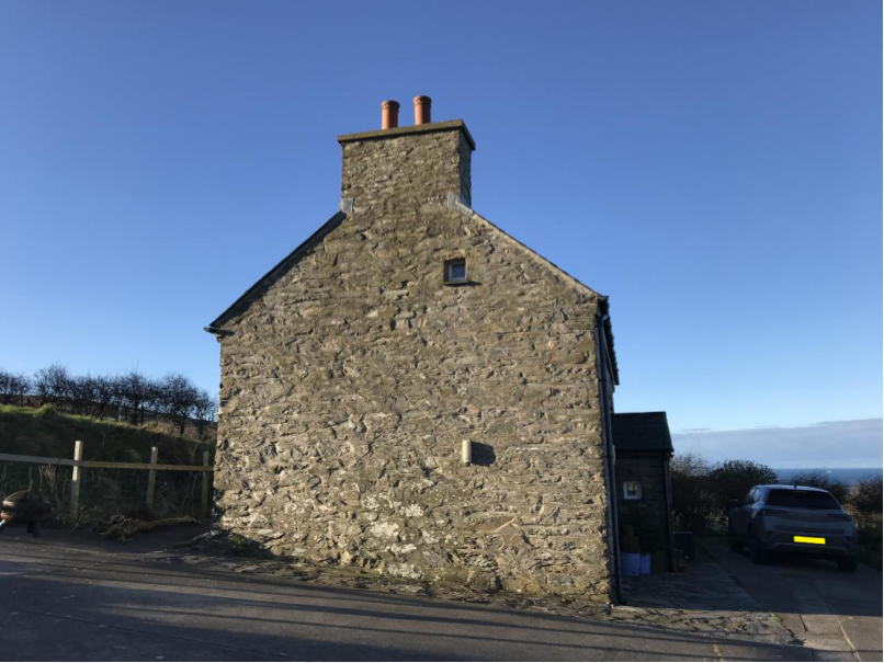 A photograph showing a detached stone building with a prominent chimney stack, situated in a rural coastal setting with a view of the sea in the background.