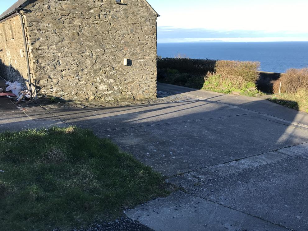 Exterior photograph of a stone building with a concrete driveway and a view of the sea in the background.