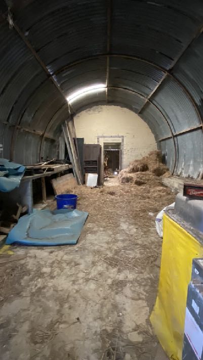 Interior photograph of a large agricultural building with a corrugated metal arched roof, containing hay and wooden structures.