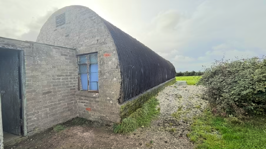 A photograph showing a long, curved-roof agricultural building, likely a Nissen hut, with a brick extension attached to the side. The structure is situated on a gravel surface in a rural setting.