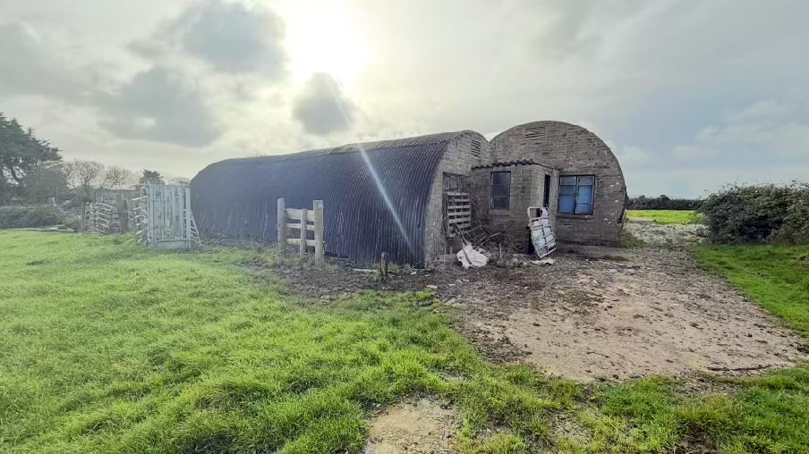 A photograph of a rural site featuring a long corrugated metal agricultural building attached to a smaller brick structure with a curved roof and a dirt track leading to it.