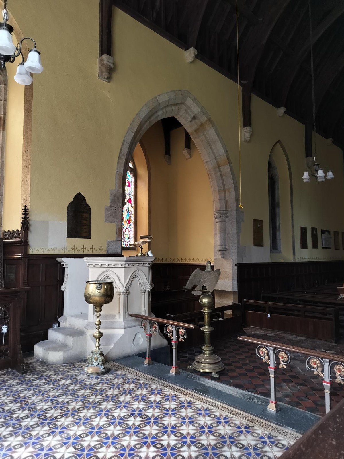 Interior view of a historic chapel featuring a stone archway, white stone font, brass vessels, and patterned tiled flooring.