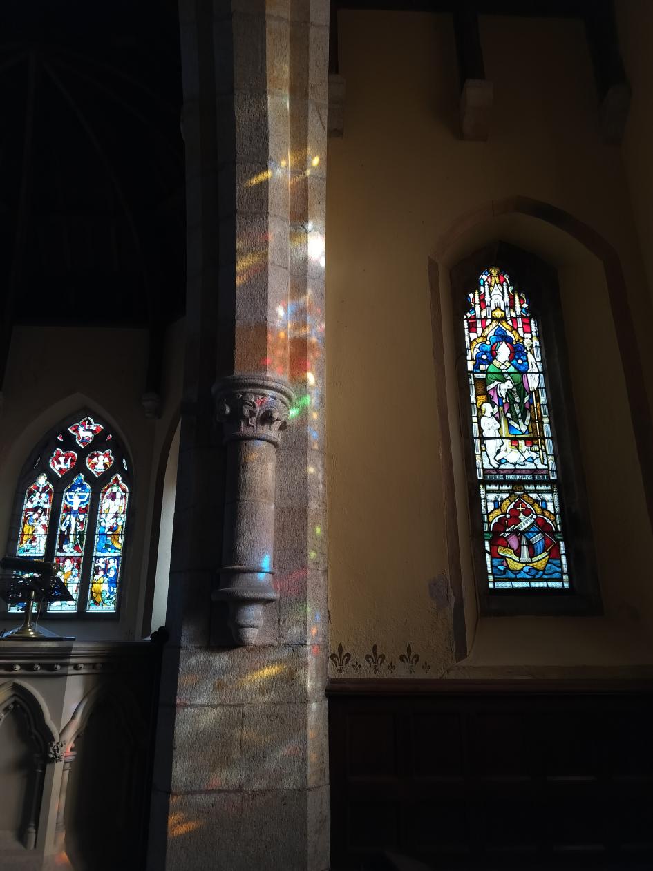 Interior photograph of a historic chapel featuring stained glass windows and a stone pillar illuminated by sunlight.
