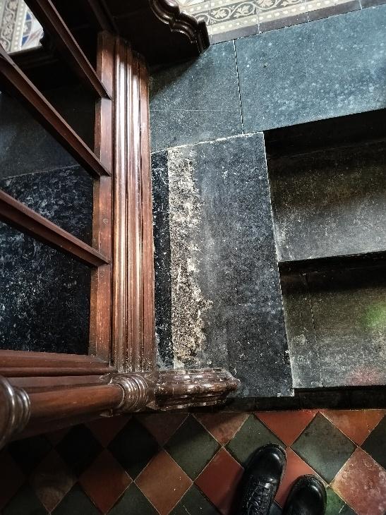 A close-up photograph of a chapel interior showing worn dark stone flooring, checkered tiles, and the leg of a wooden pew.