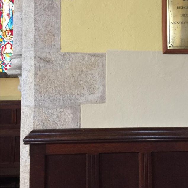 Interior photograph of a chapel showing stone masonry, a fragment of stained glass, wooden paneling, and a brass commemorative plaque.