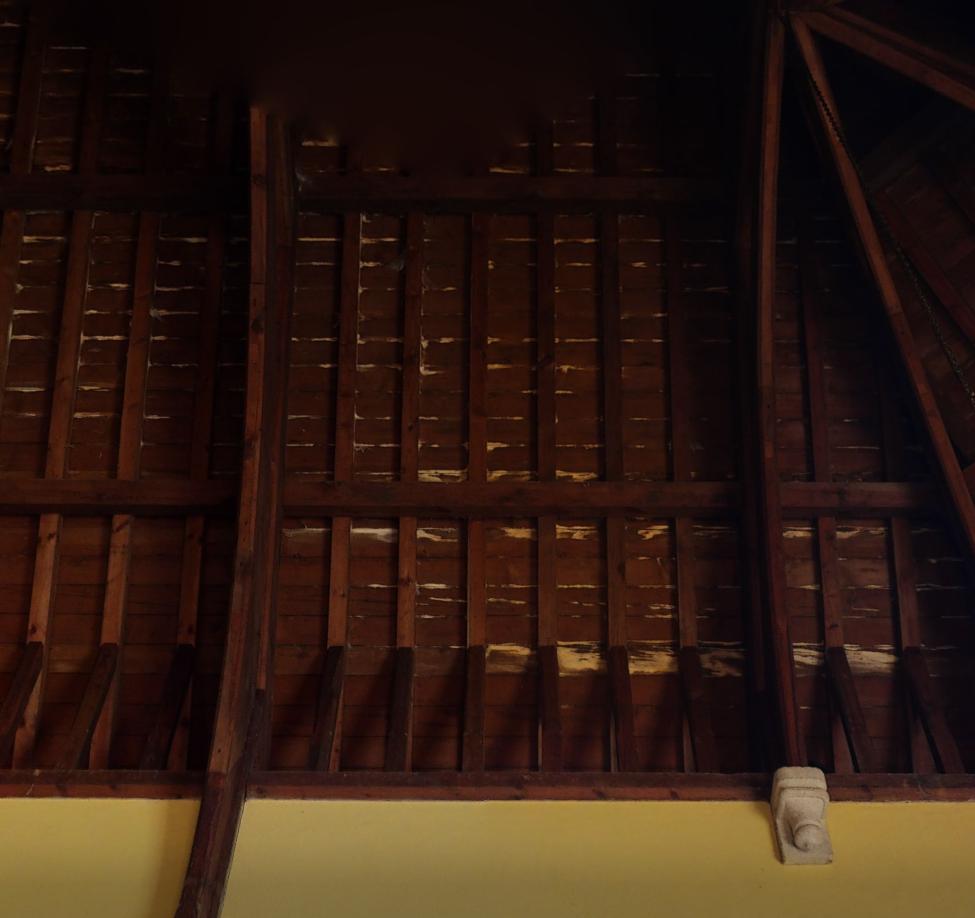 Interior photograph showing exposed wooden roof rafters and beams above a yellow wall with a stone corbel.