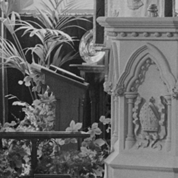 A black and white photograph showing the interior of a chapel, featuring a stone piscina or alcove with Gothic arches and a lectern surrounded by plants.