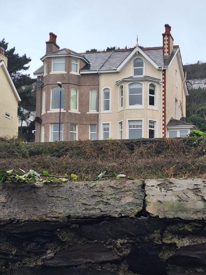 A photograph of a large detached house with mixed exterior finishes, showing brown pebbledash and cream render, viewed from behind a stone wall.