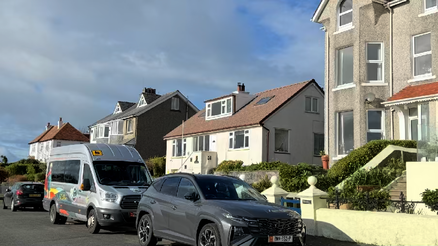 A street-level photograph showing a row of residential houses in Port Erin, with the house on the right featuring grey pebbledash rendering. Vehicles are parked on the street in the foreground.