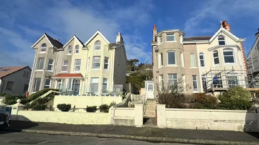 A street-level photograph showing a row of large detached houses in a coastal setting, with scaffolding erected on the right-hand property.