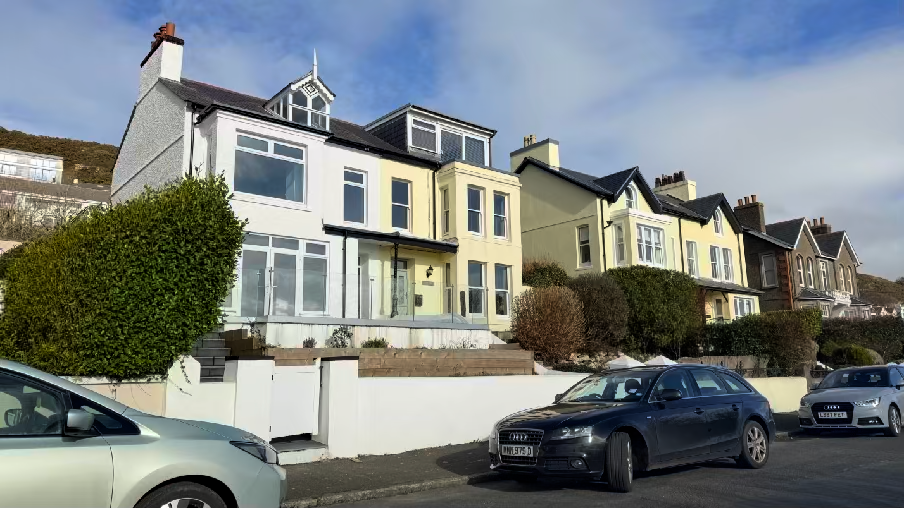 A street-level photograph showing a row of terraced houses, focusing on a white house with a large bay window and a yellow house next to it, with cars parked on the street.