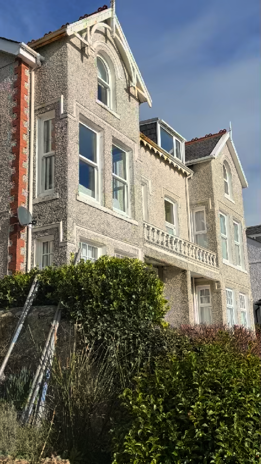 A photograph of a large, multi-story residential building featuring pebbledash rendering, white window frames, and a balcony. A ladder leans against a wall in the foreground amidst green bushes.