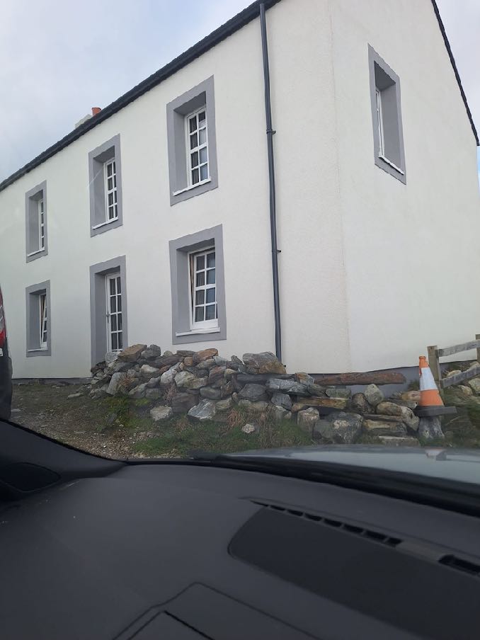A photograph taken from inside a vehicle showing the white rendered exterior of a two-story detached house with grey window surrounds and a low stone wall at the base.