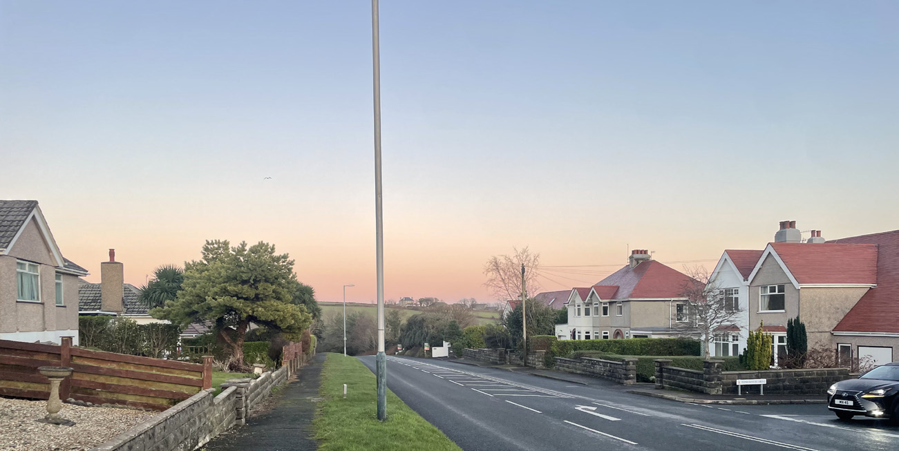 A street-level photograph showing a residential road with houses, stone walls, and hedges, looking towards open countryside in the distance.
