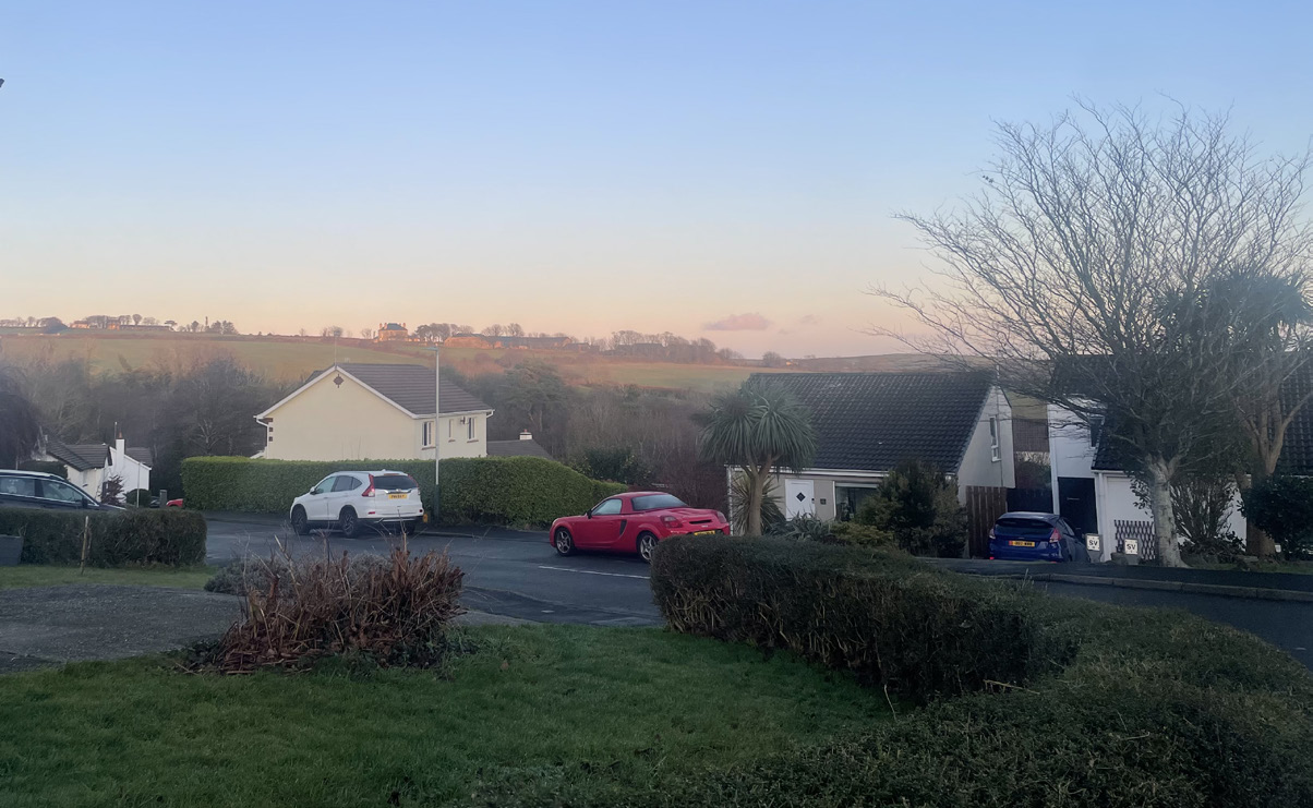 A street-level photograph showing detached houses with parked cars and a view of rolling hills in the background.