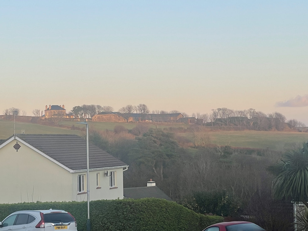 A photograph showing a rural landscape with a white house in the foreground and stone farm buildings situated on a hill in the distance.