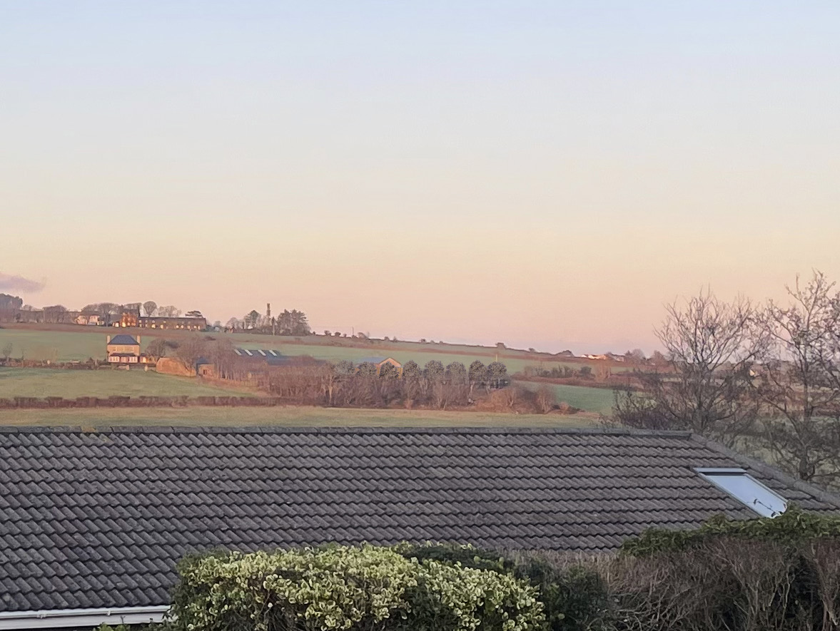 A photograph taken from a rooftop showing a grey tiled roof in the foreground and a rural landscape with fields and distant buildings in the background.