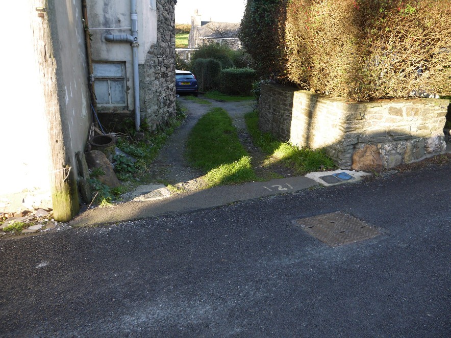 A photograph showing a narrow existing driveway or access lane between a white building and a stone boundary wall, with a car visible further up the path.
