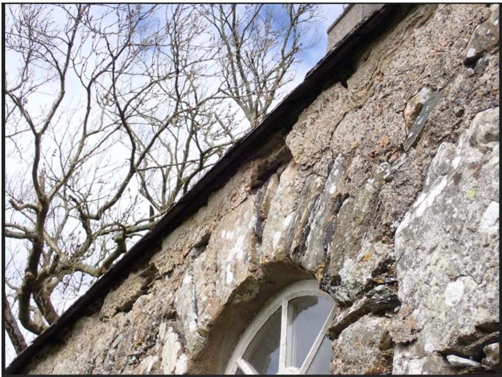 A low-angle close-up photograph showing the weathered stone masonry, roof eaves, and a white window frame of an older building against a backdrop of bare trees.