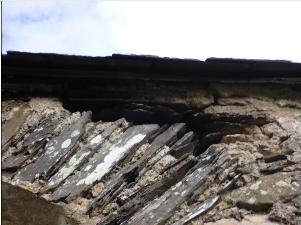 Close-up photograph of a damaged building corner showing loose slate tiles and crumbling stone masonry.