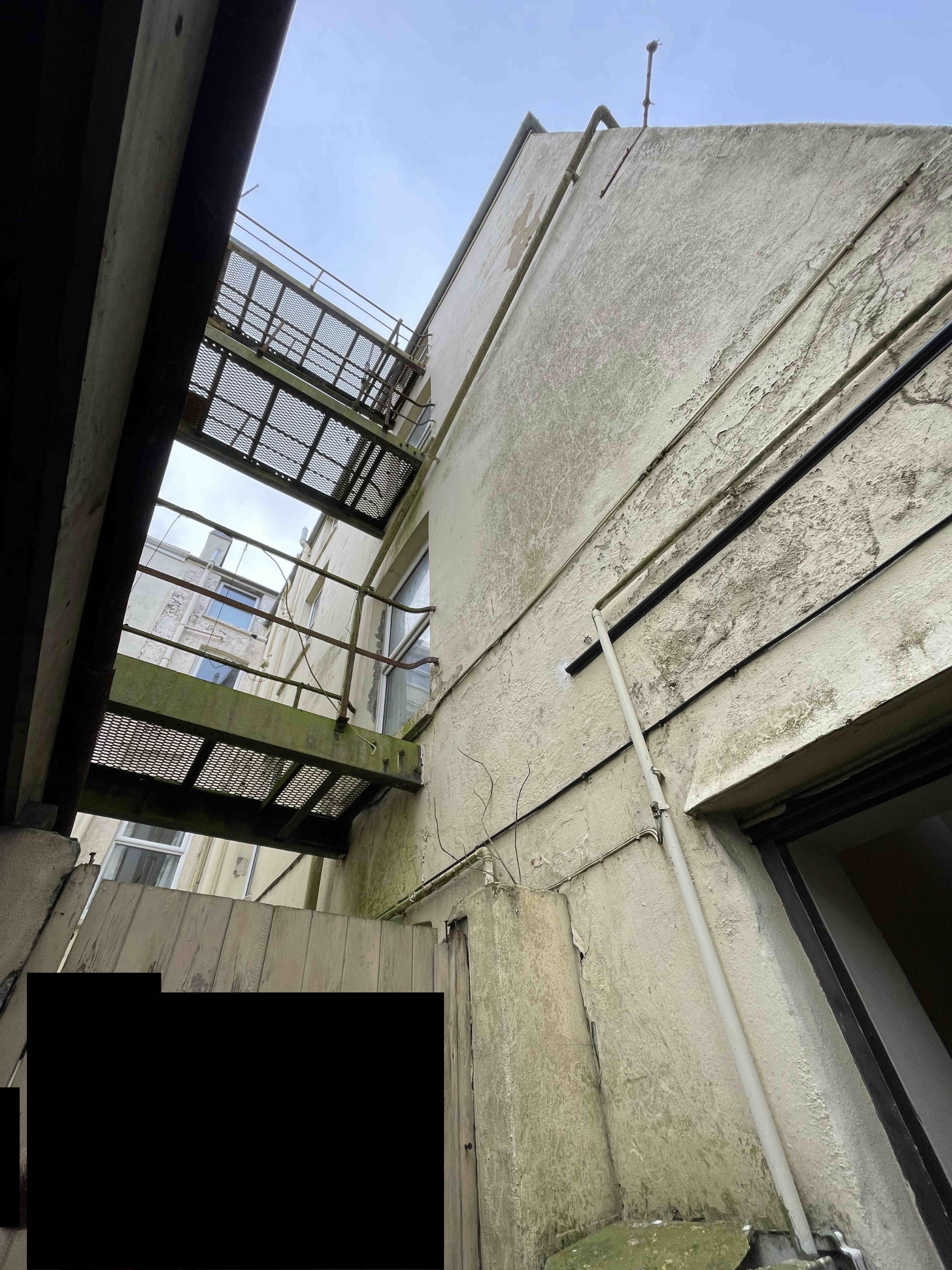 A low-angle photograph showing a weathered building exterior with a metal fire escape gantry attached to the side wall.