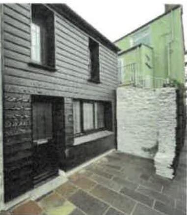 Exterior photograph showing the rear elevation of a two-story building with dark cladding, a paved yard, and an adjacent white stone wall.