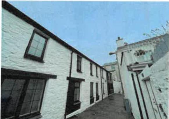 A low-angle photograph of a white-washed terraced building with dark window frames, viewed from a narrow paved rear yard or alleyway.