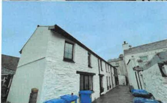 A low-angle photograph showing the side and rear elevation of a white-washed stone building, likely a terrace, with dark window frames and blue bins in the foreground.