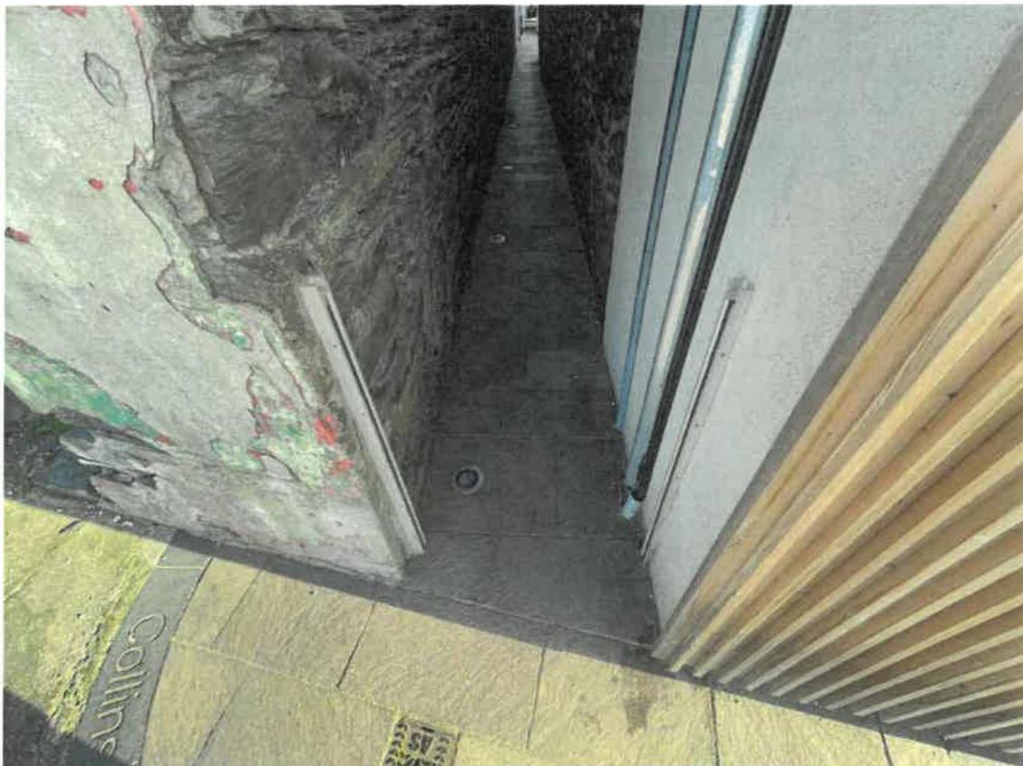 A photograph showing a narrow paved passageway or rear yard between a rough stone wall and a modern building facade with wooden slats.