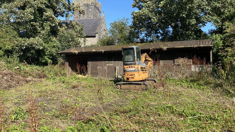 A photograph showing a dilapidated wooden stable or barn with a small yellow excavator parked in the foreground. A stone church tower is visible in the background.