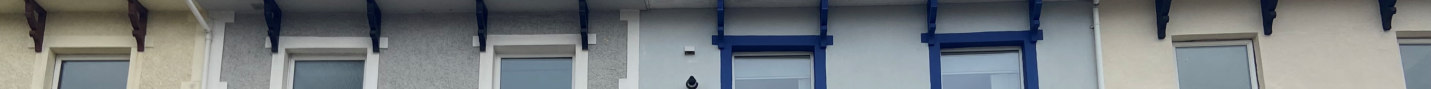 A photograph showing the upper facade of a terraced building with white and grey rendering, blue window frames, and decorative timber brackets under the eaves.