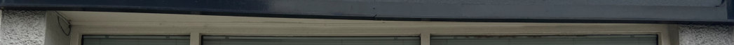A close-up photograph showing the upper section of a commercial shopfront, featuring a dark fascia board and the top of a window frame with blinds visible inside.