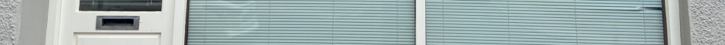 A close-up photograph showing a white door with a letterbox next to large windows covered with horizontal blinds.