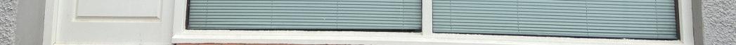 A close-up photograph of a building's ground floor exterior, showing a white door frame and a large window with horizontal blinds.