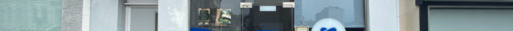 A street-level photograph showing the existing shopfront of a commercial property, featuring large glass windows, a central entrance, and a white sign with a blue heart.